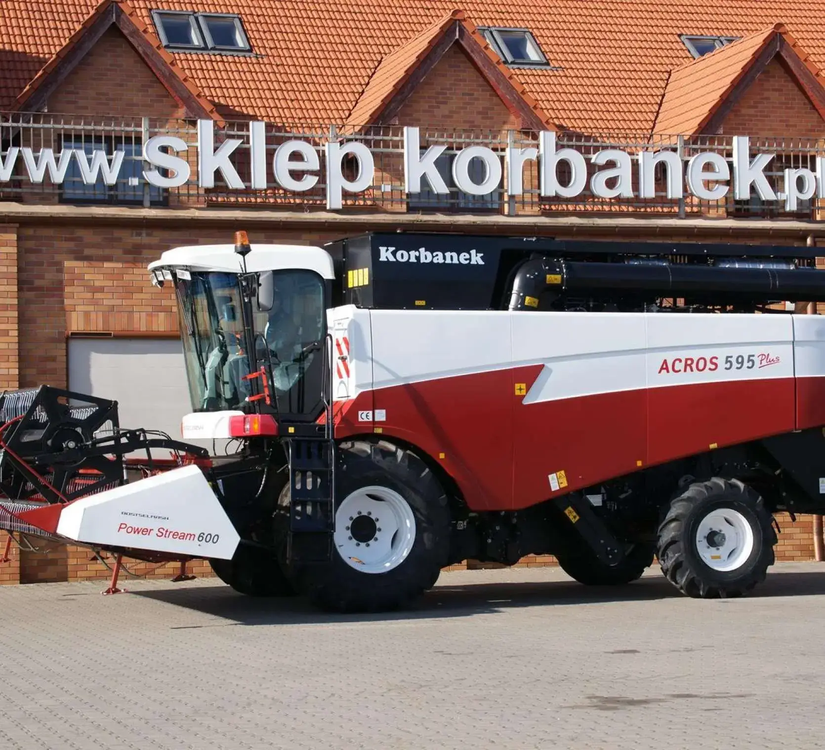 Zdjęcie boku kombajnu Acros 595+ na tle ceglanej ściany sklepu Korbanek. Kombajn Acros 595 + stojący na tle sklepu Korbanek.