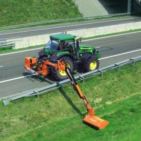 Zielony ciągnik John Deere kosi pobocze autostrady za pomocą pomarańczowej kosiarki bijakowej na wysięgniku Camel 900 firmy Samasz