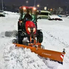 Czerwony ciągnik rolniczy z podczepionym pomarańczowym pługiem do śniegu CITY firmy Samasz odśnieża chodnik www.korbanek.pl