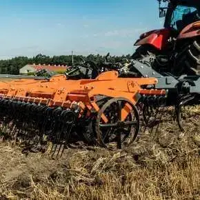 Widok z boku szaro-pomarańczowego agregatu podorywkowego KUN SUPER podczepionego do czerwonego ciągnika