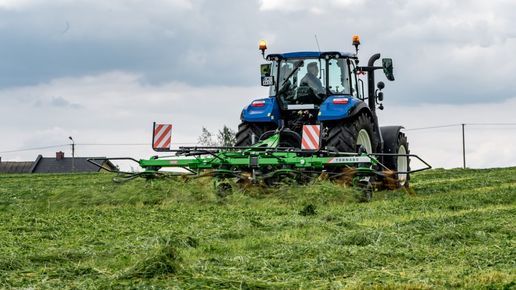 Zielony przetrząsacz TORNADO podczepiony do niebieskiego ciągnika New Holland podczas pracy na łące