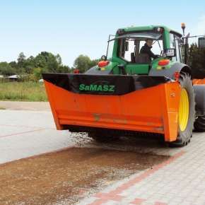 Widok tylny zielonego ciągnika rolniczego z zawieszoną posypywarką SAHARA 240 podczas sypania piasku na placu www.korbanek.pl