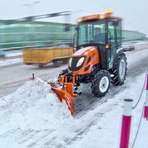 Pomarańczowy ciągnik rolniczy z zaczepionym pomarańczowym pługiem do śniegu CITY 180 firmy Samasz odśnieża chodnik www.korbanek.pl