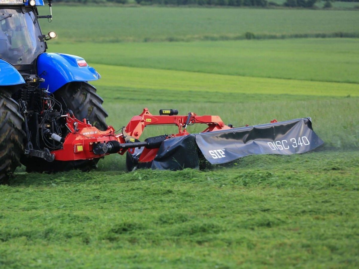 Niebieski traktor rolniczy z czerwoną tylną kosiarką dyskową SILVERCUT DISC 340S podczas pracy