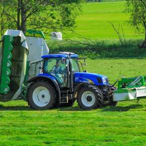 Niebieski ciągnik rolniczy New Holland jedzie z kosiarką dyskową do trawy MegaCUT po łące www.korbanek.pl